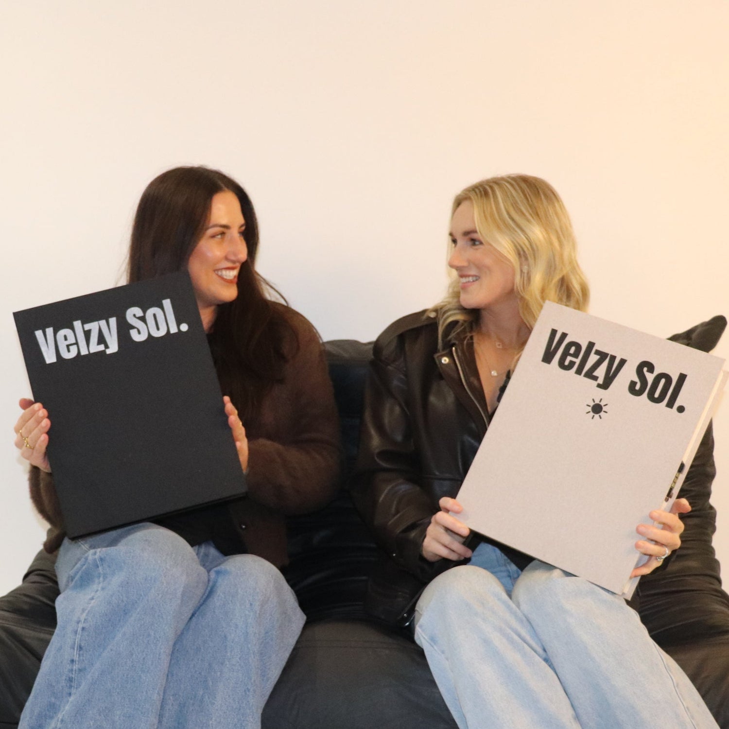 Two women sitting on a black couch holding 'Velzy Sol.' book box products against a white background